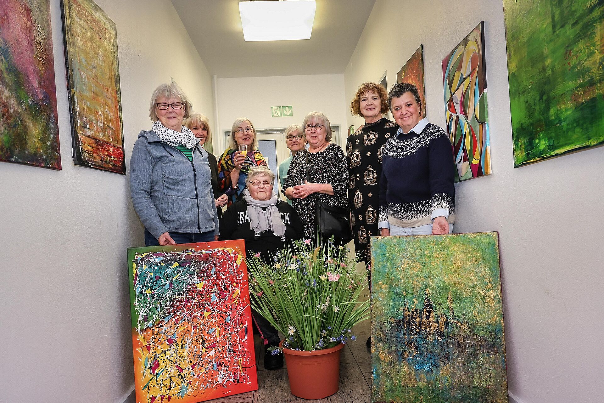 Mehere Frauen stehen im Gang der AWO-Begegnungsstätte, lächeln in die Kamera und zeigen bei der Ausstellungseröffnung ihre gemalten Bilder. 