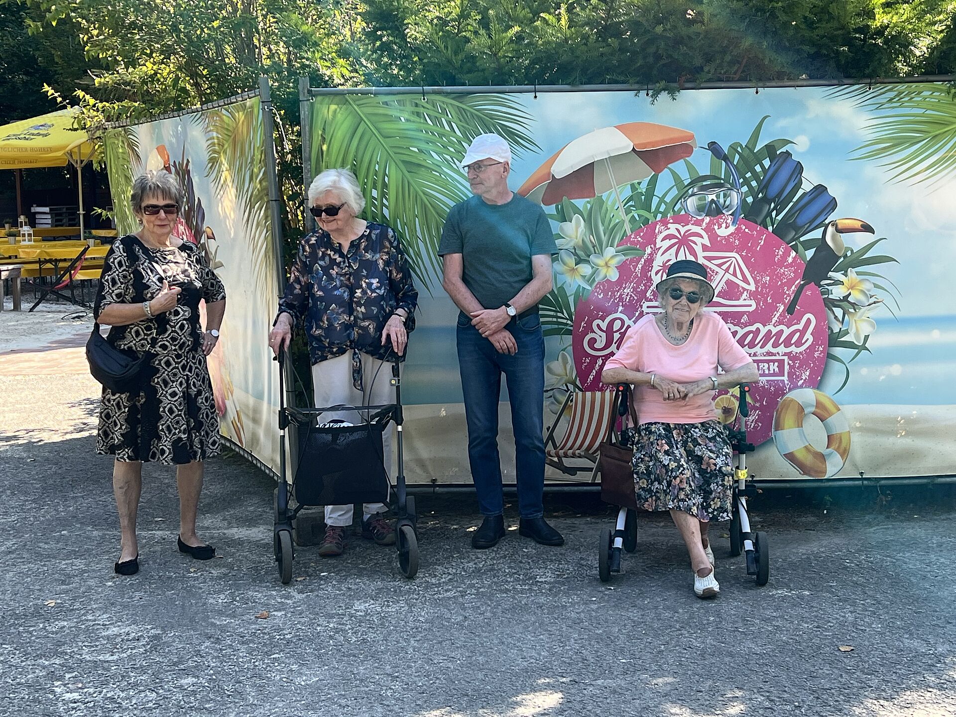 Drei Seniorinnen und ein Senior stehen im Westfalenpark vor dem bunten Banner eines Cafés.