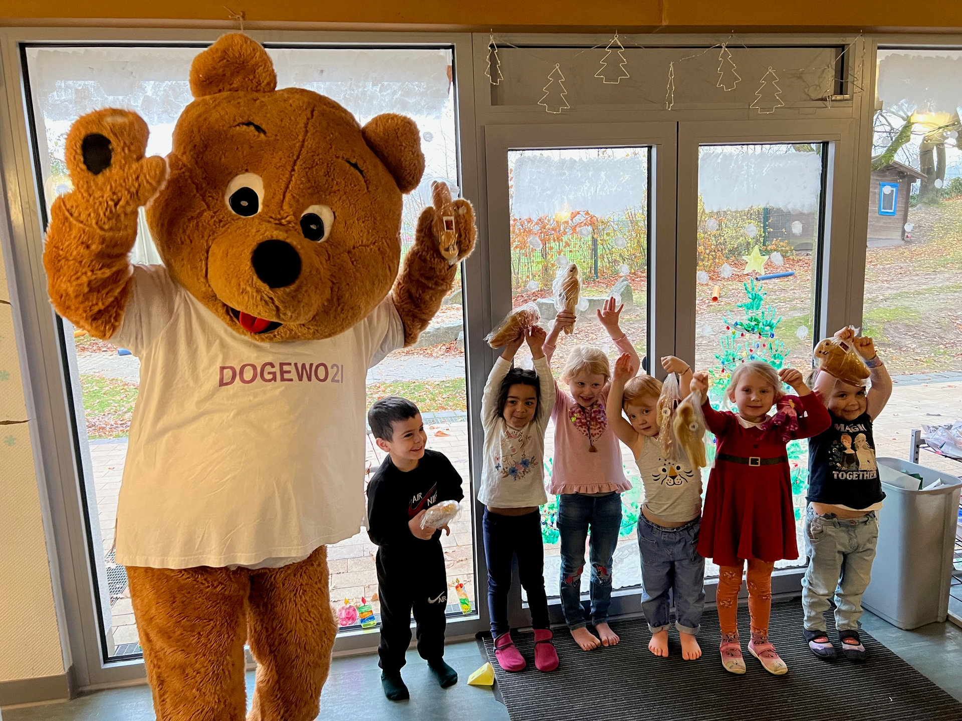 Ein Bärenmaskottchen, der Dogibär, und Kinder lachen in die Kamera und zeigen ihre Stutenkerle