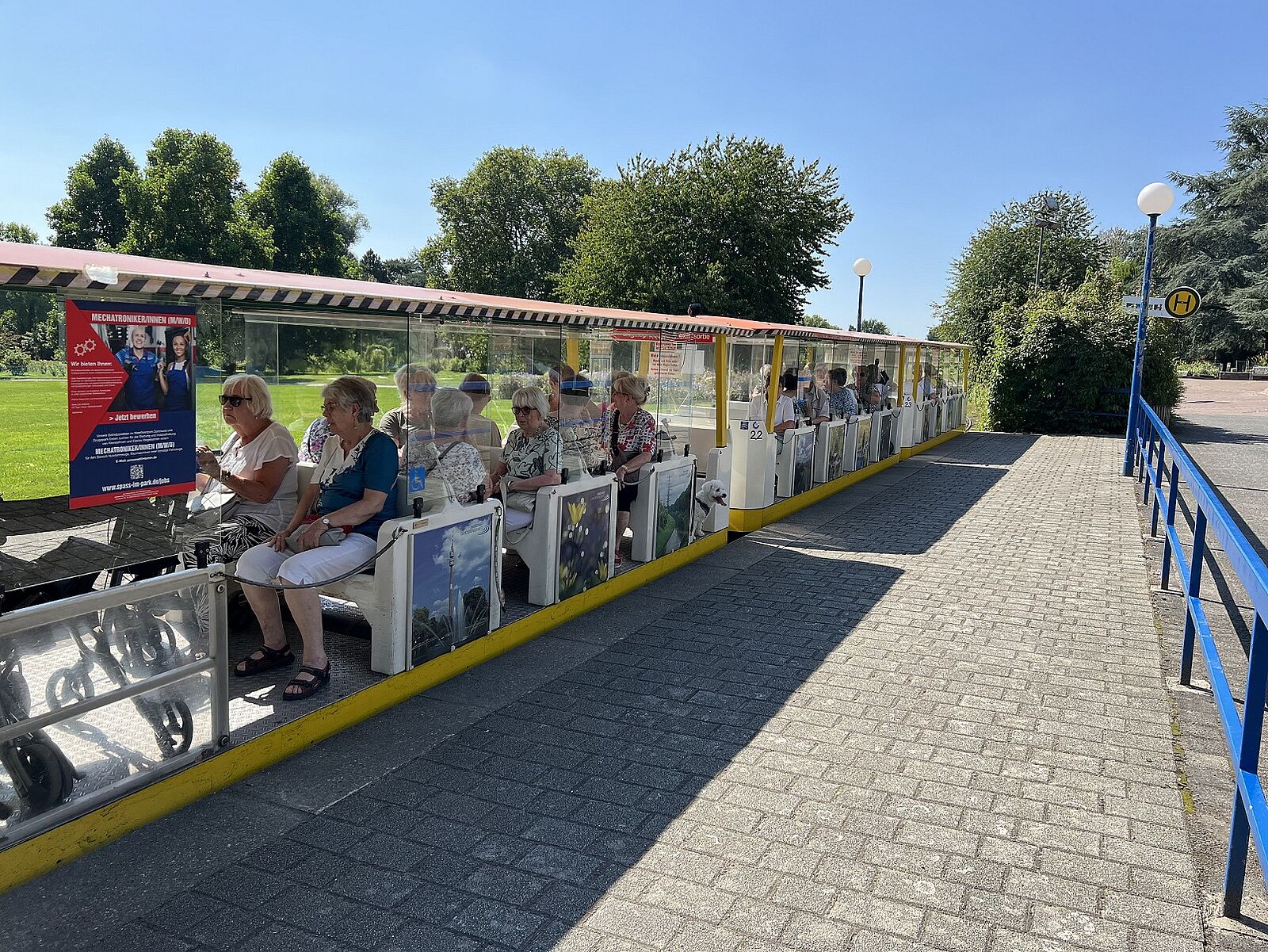 Frauen und Männer sitzen bei gutem Wetter gemeinsam mit dem kleinen, weißen Hund Max in der Kleinbahn des Westfalenparks.